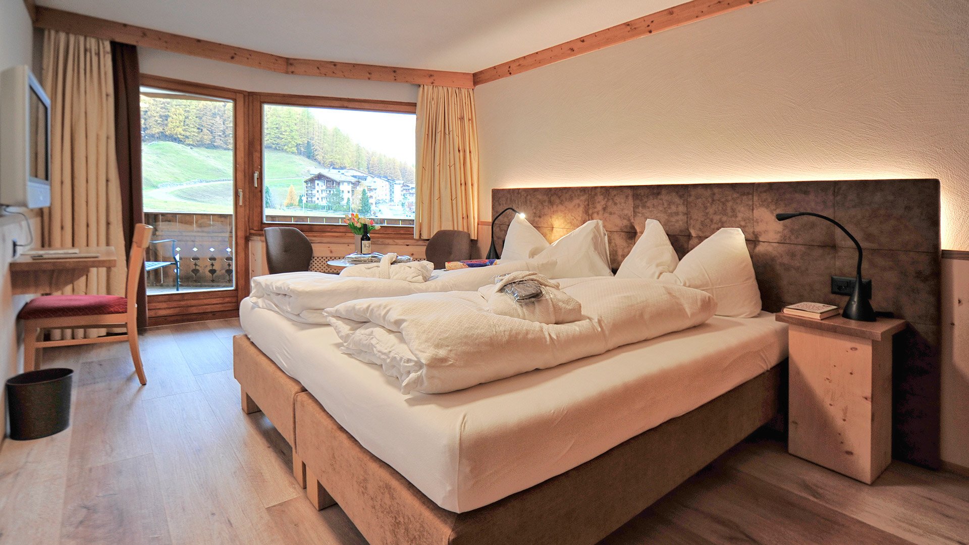 Gemütliches Hotelzimmer mit Doppelbett, Holzmöbeln und Aussicht auf grüne Hänge und Chalets durch großes Fenster.
