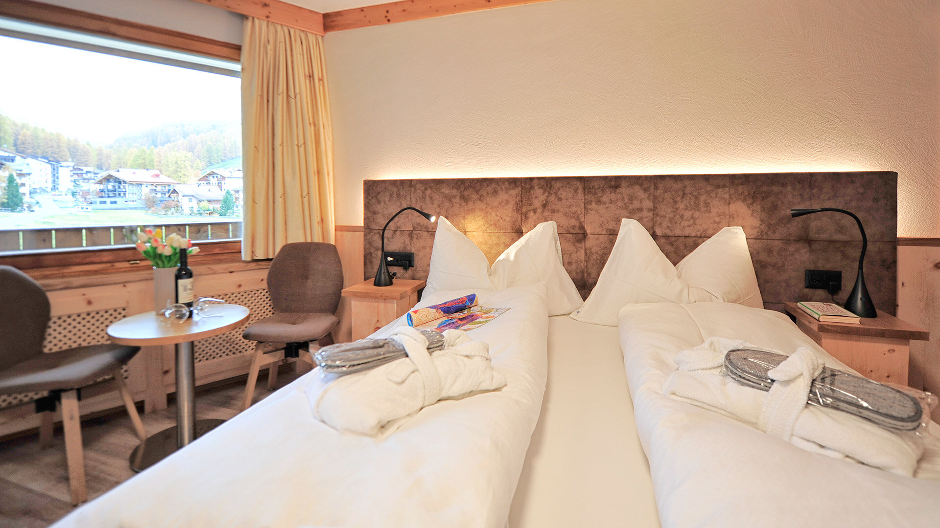 Gemütliches Hotelzimmer mit Doppelbett, Holzmöbeln und Aussicht auf grüne Hänge und Chalets durch großes Fenster.