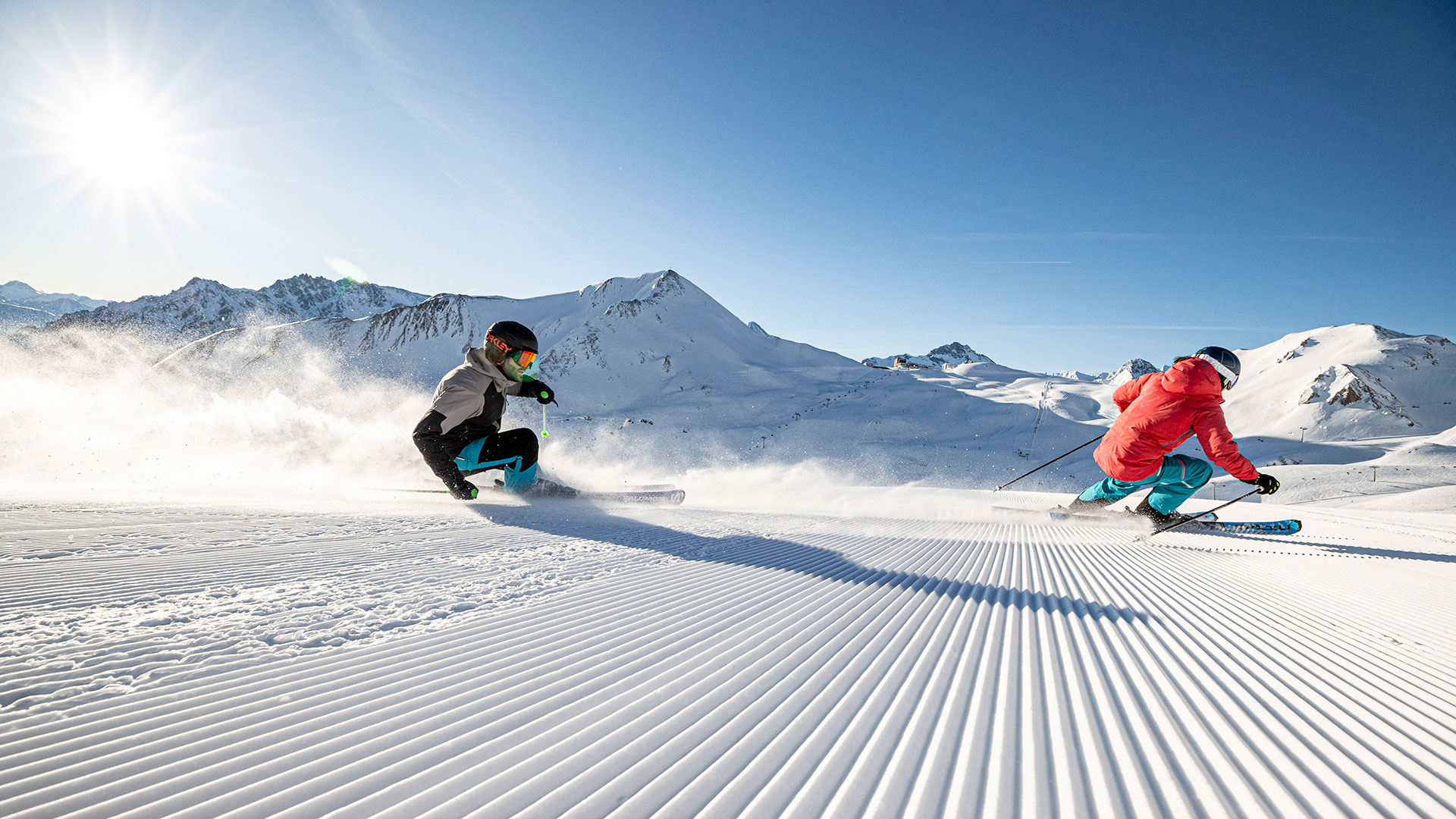 Zwei Skifahrer carven bei strahlendem Sonnenschein auf frisch präparierter Piste in einer verschneiten Berglandschaft.