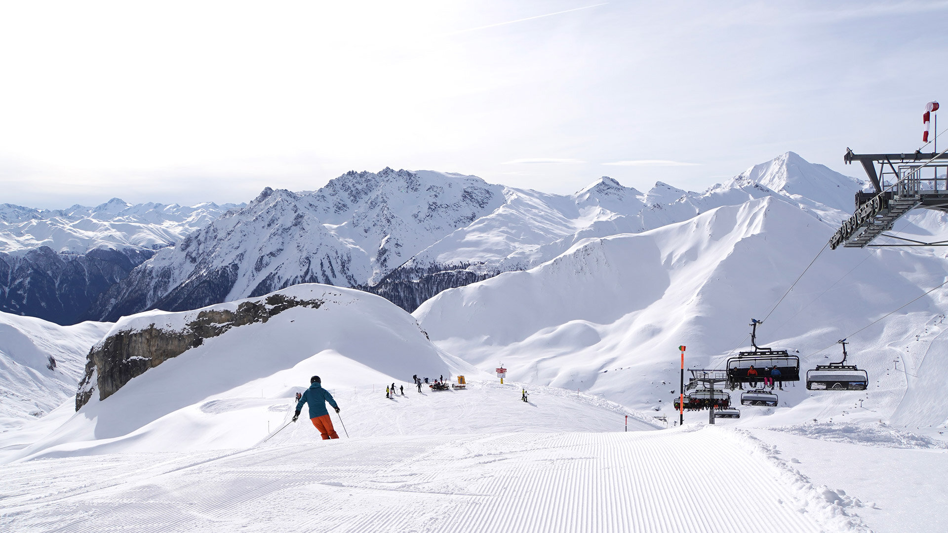 Skifahrer fährt Piste hinab, im Hintergrund schneebedeckte Berge und Sessellift unter leicht bewölktem Himmel.